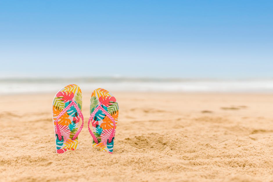 Vibrant patterned flip flops standing on a sandy beach with a clear blue sky.